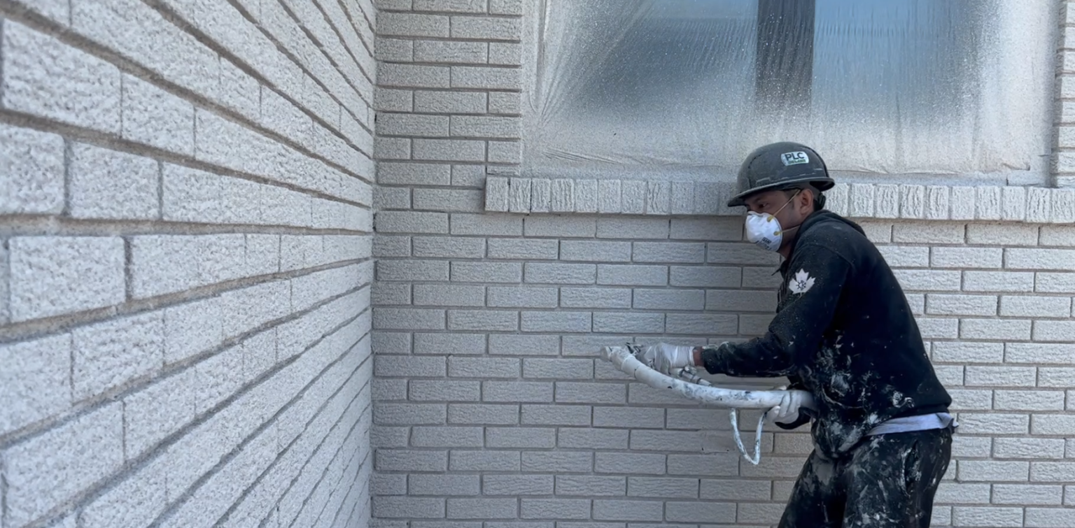 A man wearing a black jacket and hat stands beside a house applying cork spray.