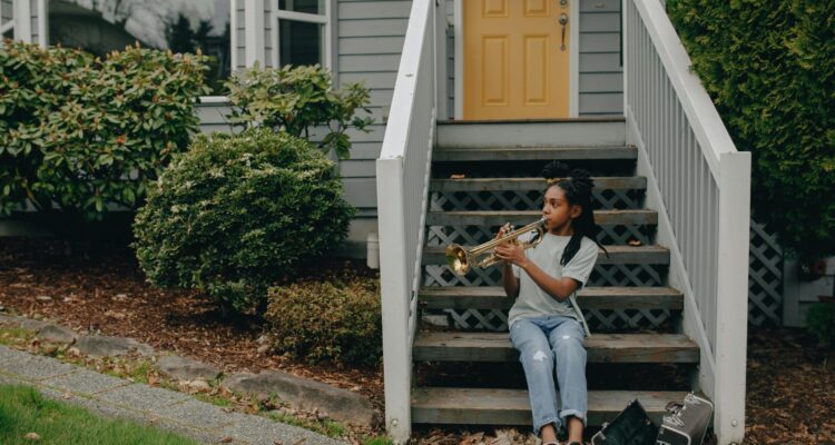 A young girl sits on the steps of her house playing a trumpet, smiling and enjoying a sunny day in her neighborhood.