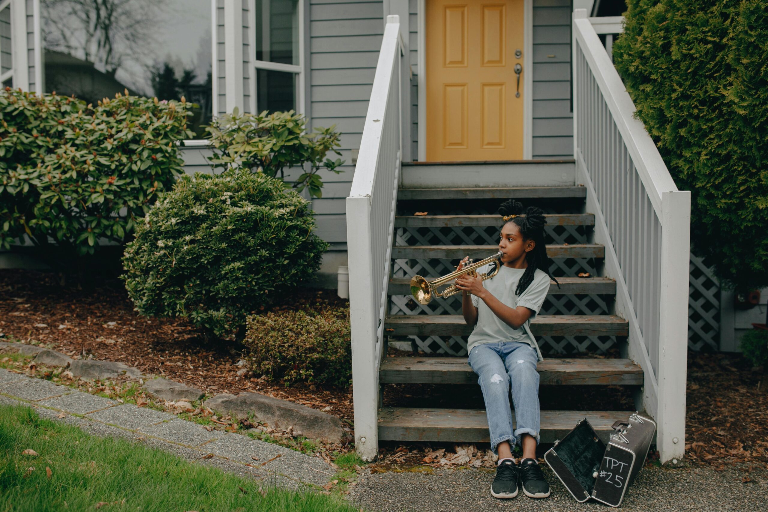 A young girl sits on the steps of her house playing a trumpet, smiling and enjoying a sunny day in her neighborhood.