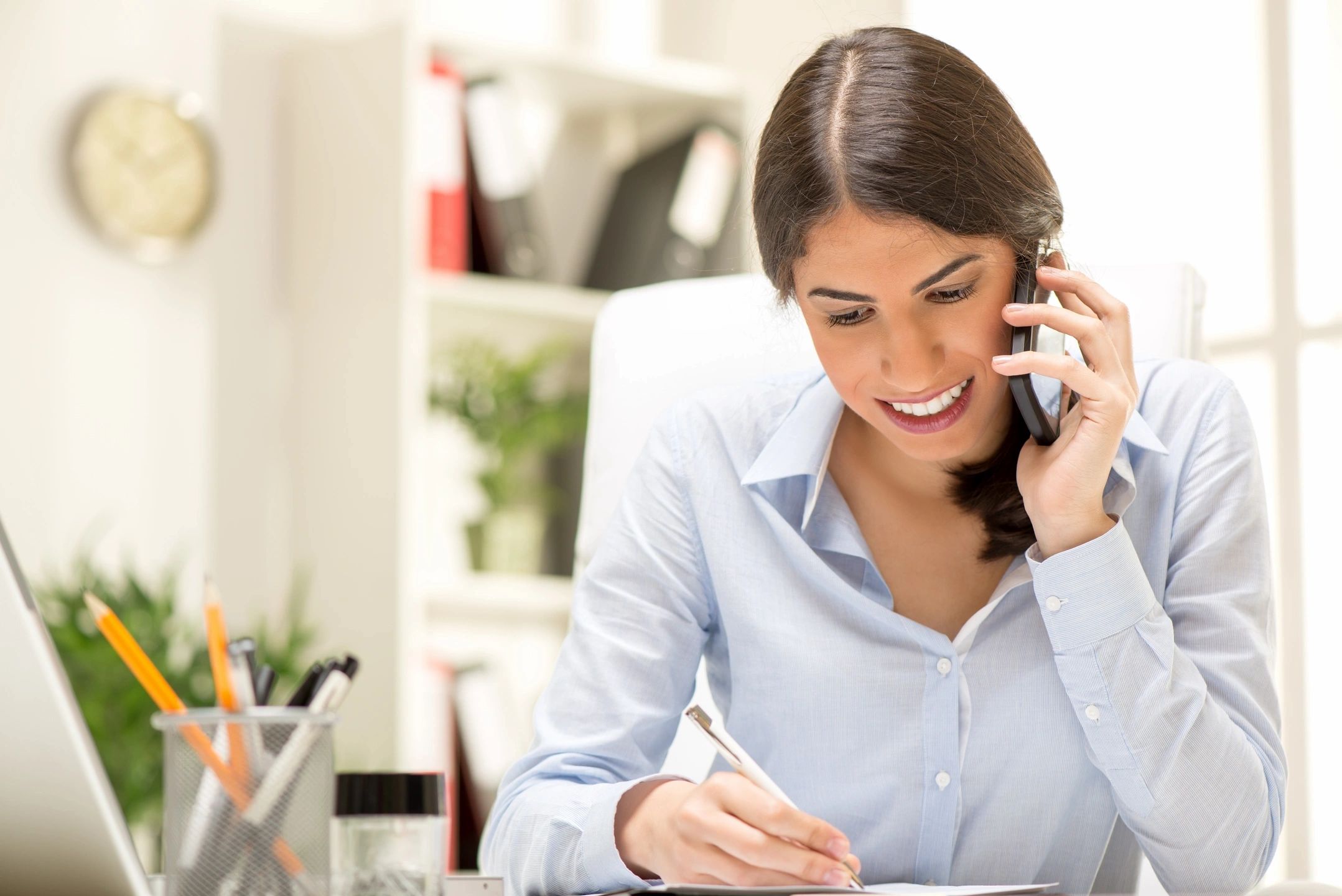 A woman seated at a desk, working on a laptop while holding a phone in her hand.